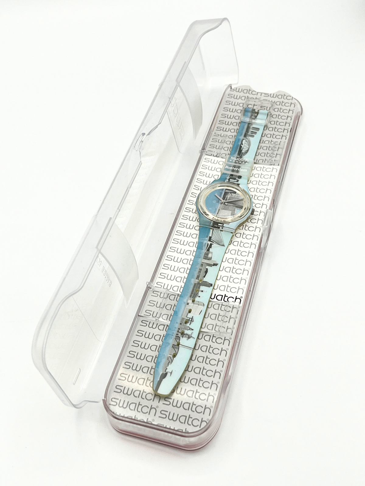 A Swatch wristwatch with a light blue patterned strap and silver-toned case, presented inside its clear plastic protective packaging on a white background.