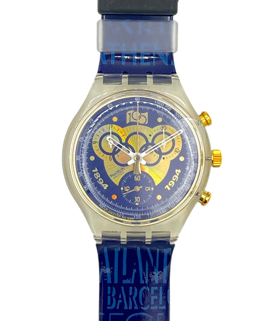 A Swatch wristwatch featuring a blue strap with 'ATLANTA BARCELONA FEST' text, a gold-tone case, and a blue dial with a yellow and white 1994 Olympic rings graphic.