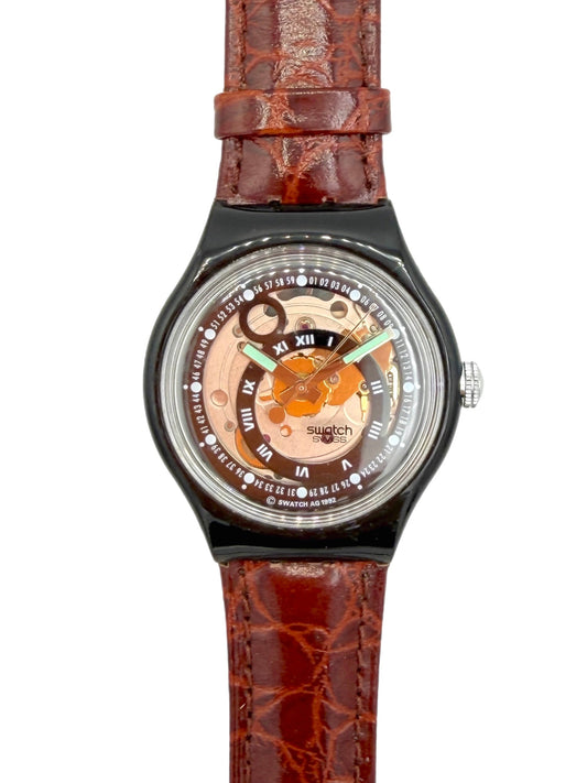A wristwatch with a brown textured leather strap, a black bezel, and a transparent dial showing a gold-toned inner mechanism and Roman numeral hour markers.
